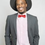 Photo of Jarell smiling wearing a gray hat and jacket with a pink shirt and bowtie.